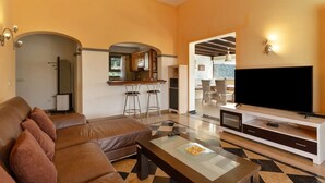 Un salon avec un canapé en cuir marron, une table basse en verre, un téléviseur à écran plat posé sur un meuble de télévision blanc, et un coin cuisine avec un bar et des tabourets.