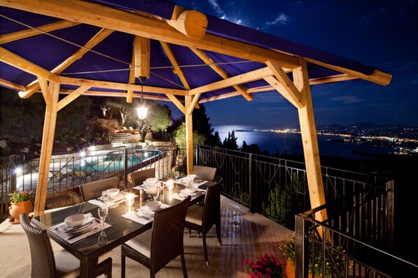 A covered outdoor dining area with a pool and city lights in the background.