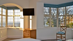A bedroom with a large bed, a wooden dresser, a chair, a television, and large windows with a view of trees and buildings. - Large house with sea views, ample off-road parking, enclosed garden with terrace (Hunstanton)