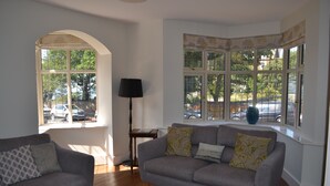 A living room with a grey sofa and armchair, a lamp, and large windows with views of trees and parked cars. - Large house with sea views, ample off-road parking, enclosed garden with terrace (Hunstanton)