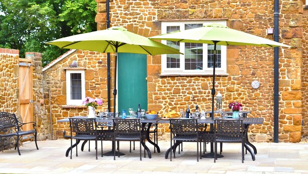 An outdoor patio with a stone wall, a table set with chairs, and green umbrellas. - Large house with sea views, ample off-road parking, enclosed garden with terrace (Hunstanton)