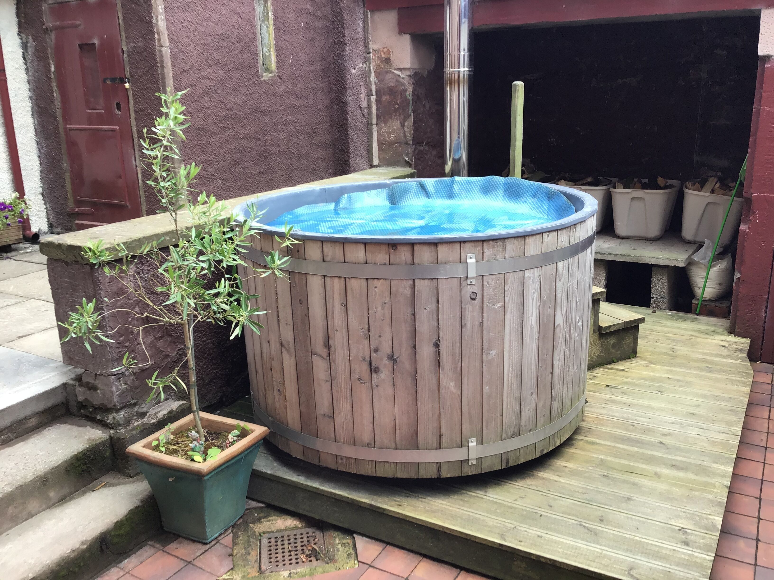 Un bain à remous en bois, avec un intérieur bleu, entouré d’une terrasse en bois et agrémenté d’une plante en pot.