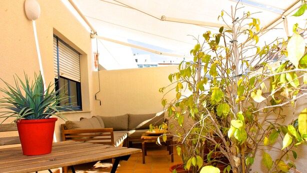 Ein Terrassenbereich im Freien mit einem Holztisch, roten und braunen Blumentöpfen, einer Holzbank und einer Couch.