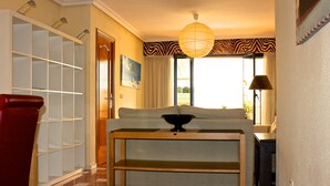 Ein Hotelzimmer mit einer Couch, einem Holztisch mit einer Schüssel, einem roten Sessel, einem Bücherregal und einem Fenster mit Vorhängen.
