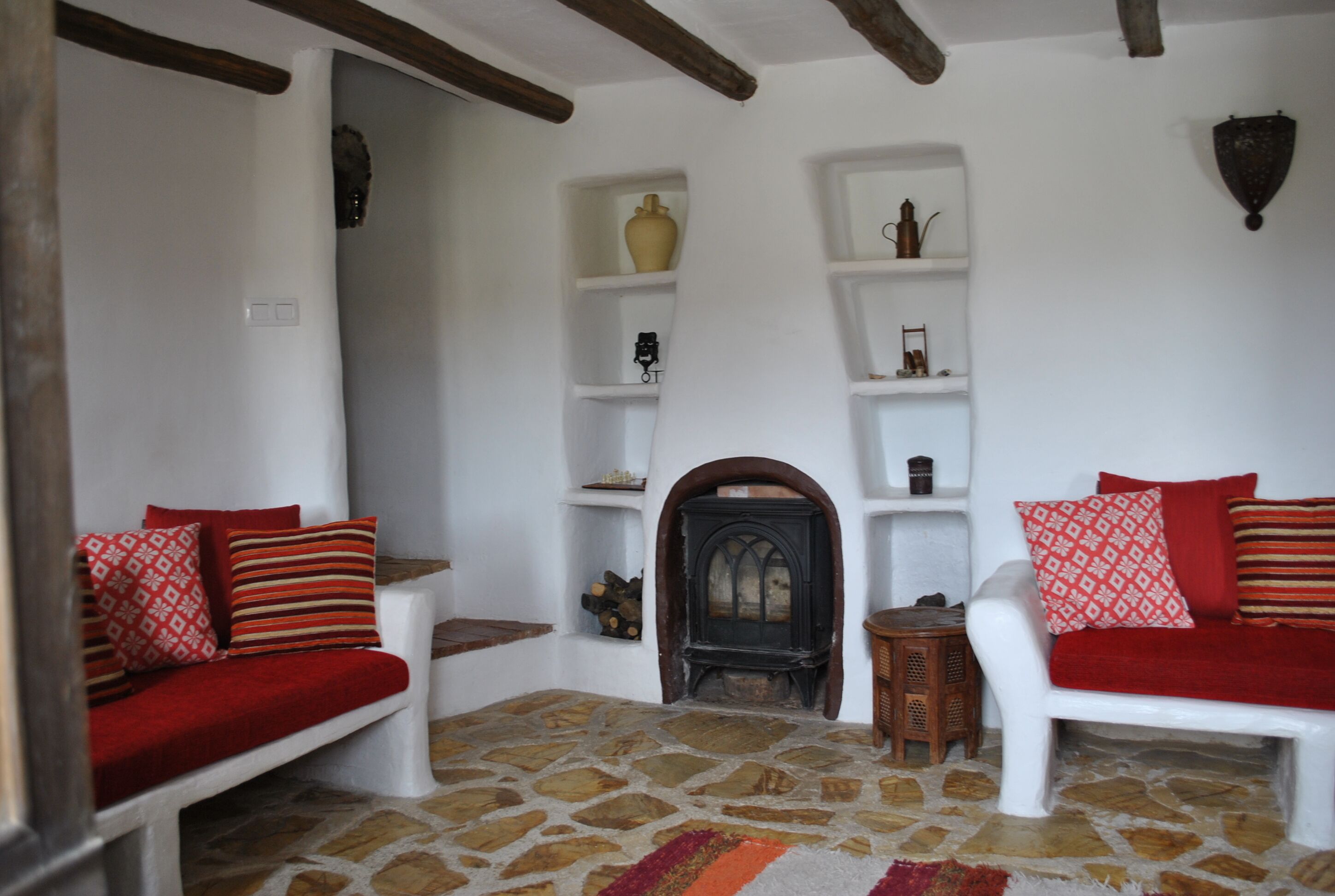 Una habitación acogedora con suelo de piedra, paredes blancas y vigas de madera. Hay dos sofás blancos con cojines rojos, una mesita de madera y una estantería con varios objetos decorativos.