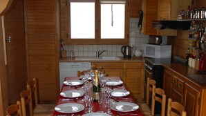 Een eetgedeelte met een rode tafel gedekt voor een maaltijd, houten stoelen en een keuken op de achtergrond.
