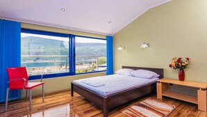 Une chambre à coucher avec un lit en bois, une chaise rouge, une table de chevet, un vase de fleurs et une vue sur le paysage urbain à travers de grandes fenêtres.