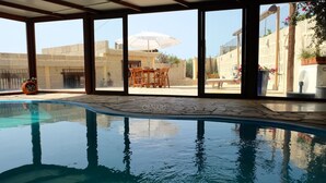 Piscina interior com paredes de vidro transparente, chão ladrilhado e vista para um pátio externo com mesa e guarda-sol.