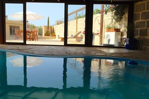 Ein Innenpool mit großen Glaswänden, gefliestem Boden und Blick auf eine Außenpatio-Terrasse mit Tisch und Stühlen.