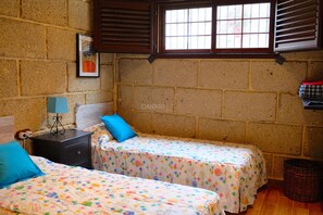 A room with two beds, a nightstand, a lamp, a window with shutters, a wall-mounted picture, and a basket. - Charming Two-Bedroom Country House with Heated Pool (Arico)