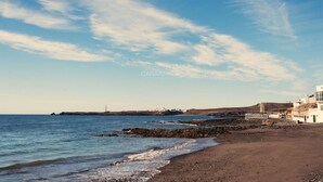 Uma praia costeira com um trecho rochoso, um farol à distância e um edifício branco com varanda com vista para o mar.