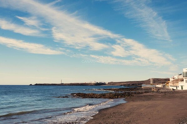 Une plage côtière bordée d’un rivage rocheux, un phare visible au loin et un bâtiment blanc doté d’un balcon donnant sur la mer.