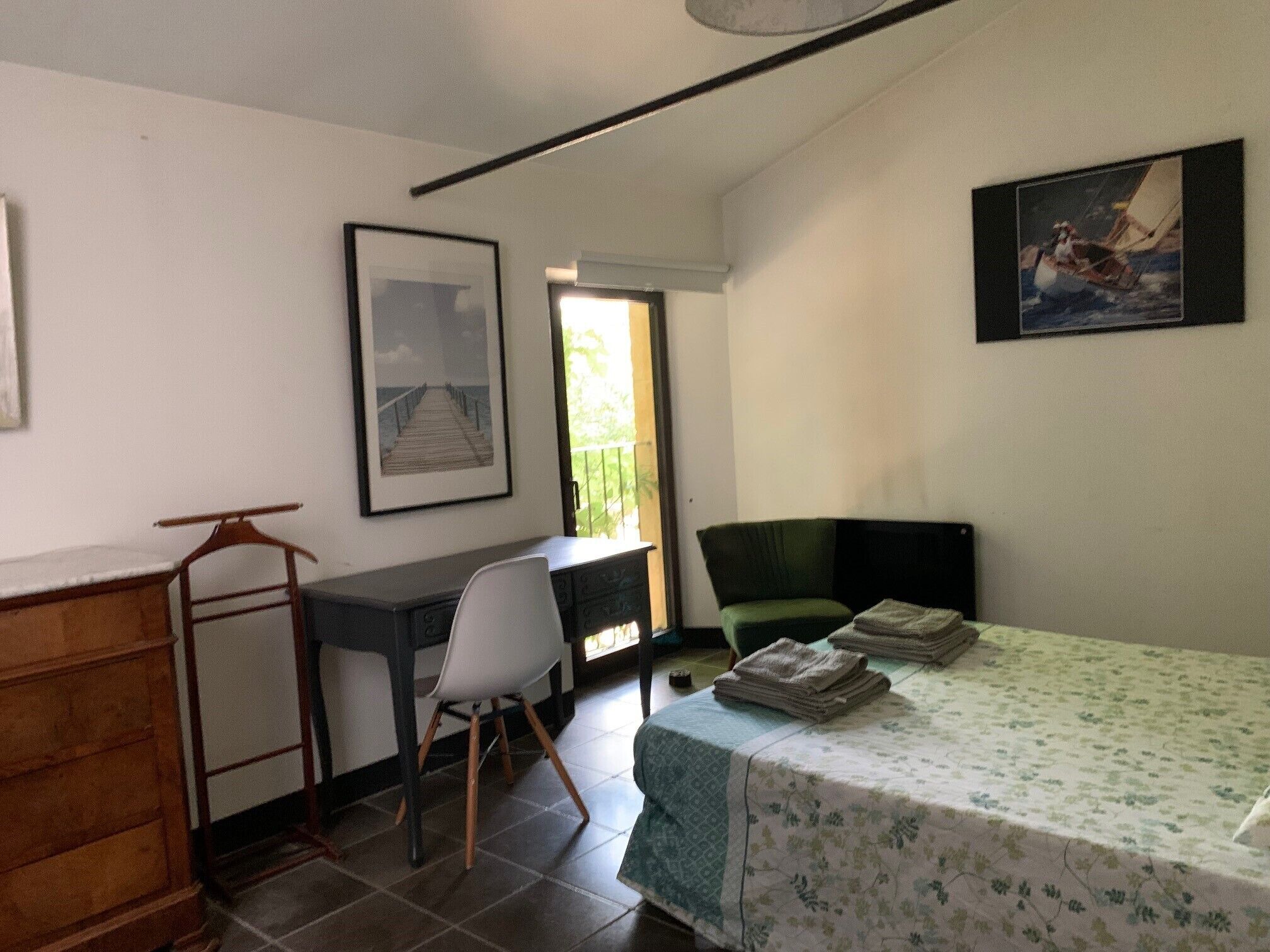 A small, tidy room with a desk, a chair, a bed with a floral bedspread, a green armchair, and two framed pictures on the walls.