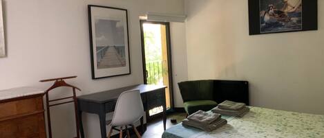 A small, tidy room with a desk, a chair, a bed with a floral bedspread, a green armchair, and two framed pictures on the walls.
