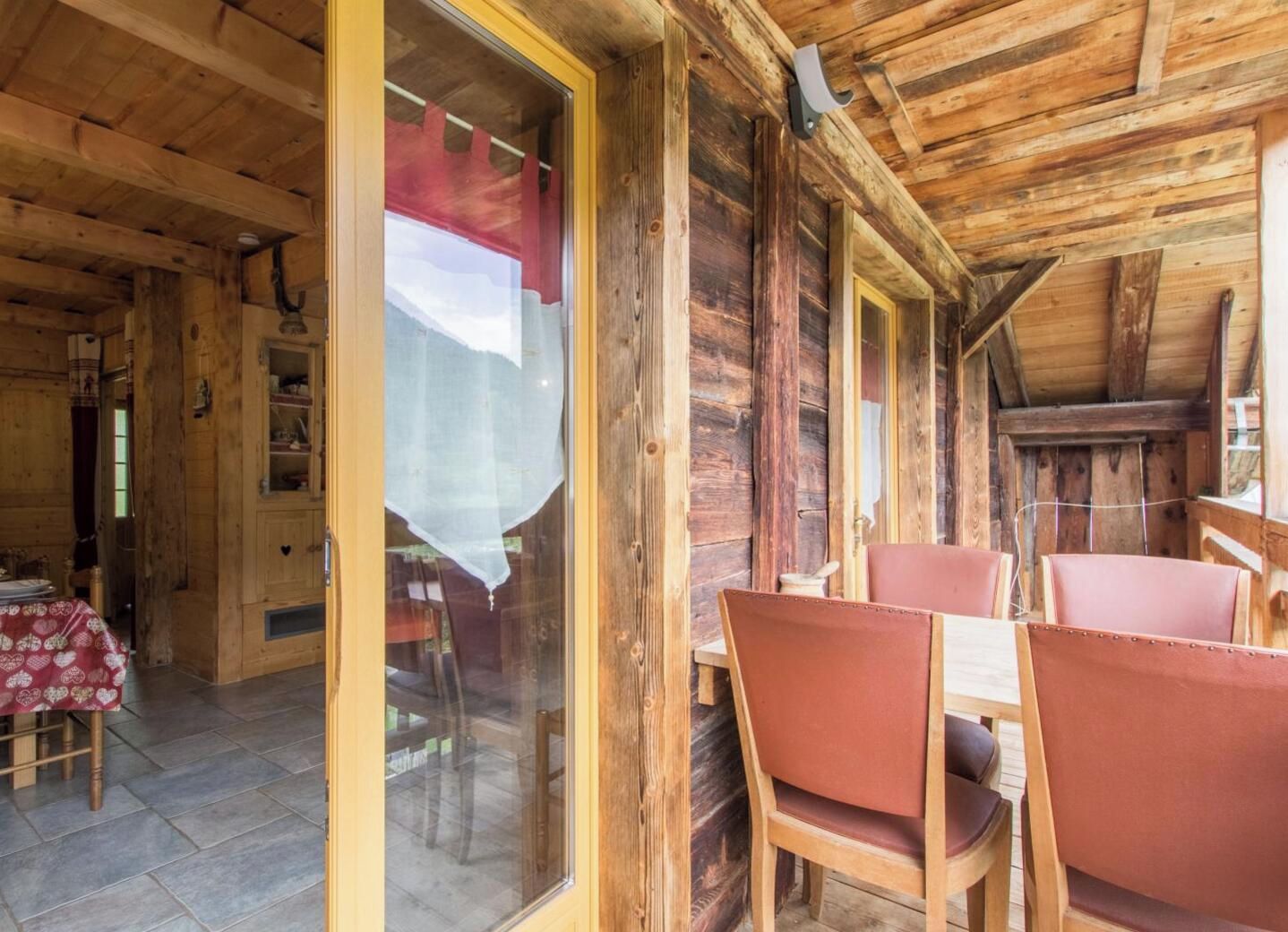A wooden interior with a dining area featuring tables and chairs, a visible kitchen area with cabinets, and a large window with a curtain.