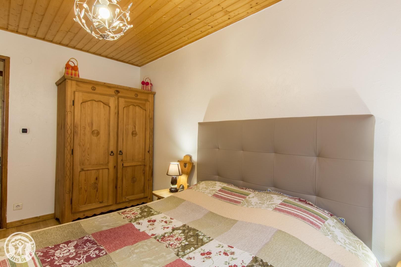 Une chambre à coucher avec une armoire en bois, un lit doté d’une tête de lit rembourrée de couleur grise et une courtepointe ornée de motifs floraux.