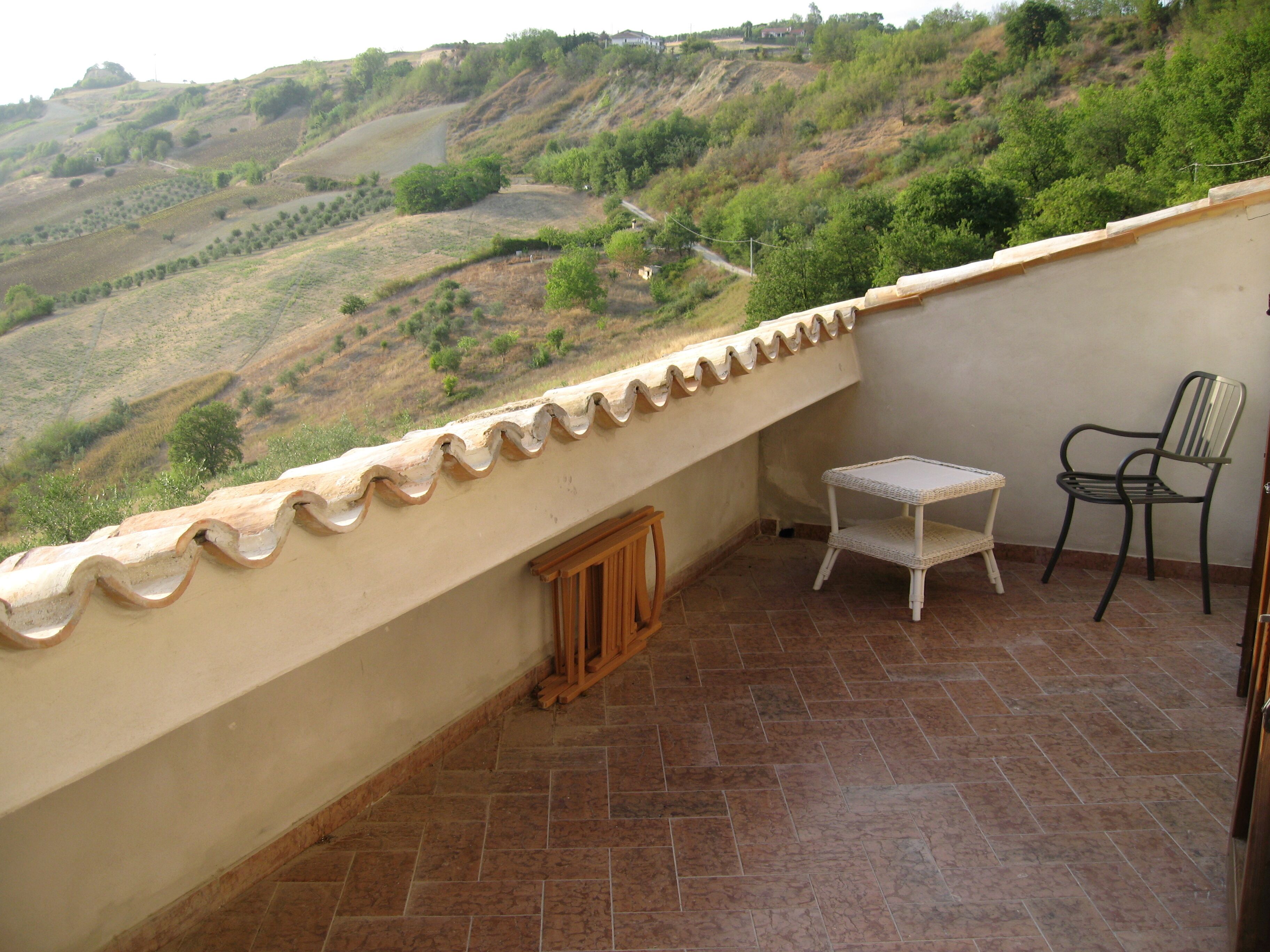 A balcony with a view of a hilly landscape, a small table, a chair, and a wooden cabinet.