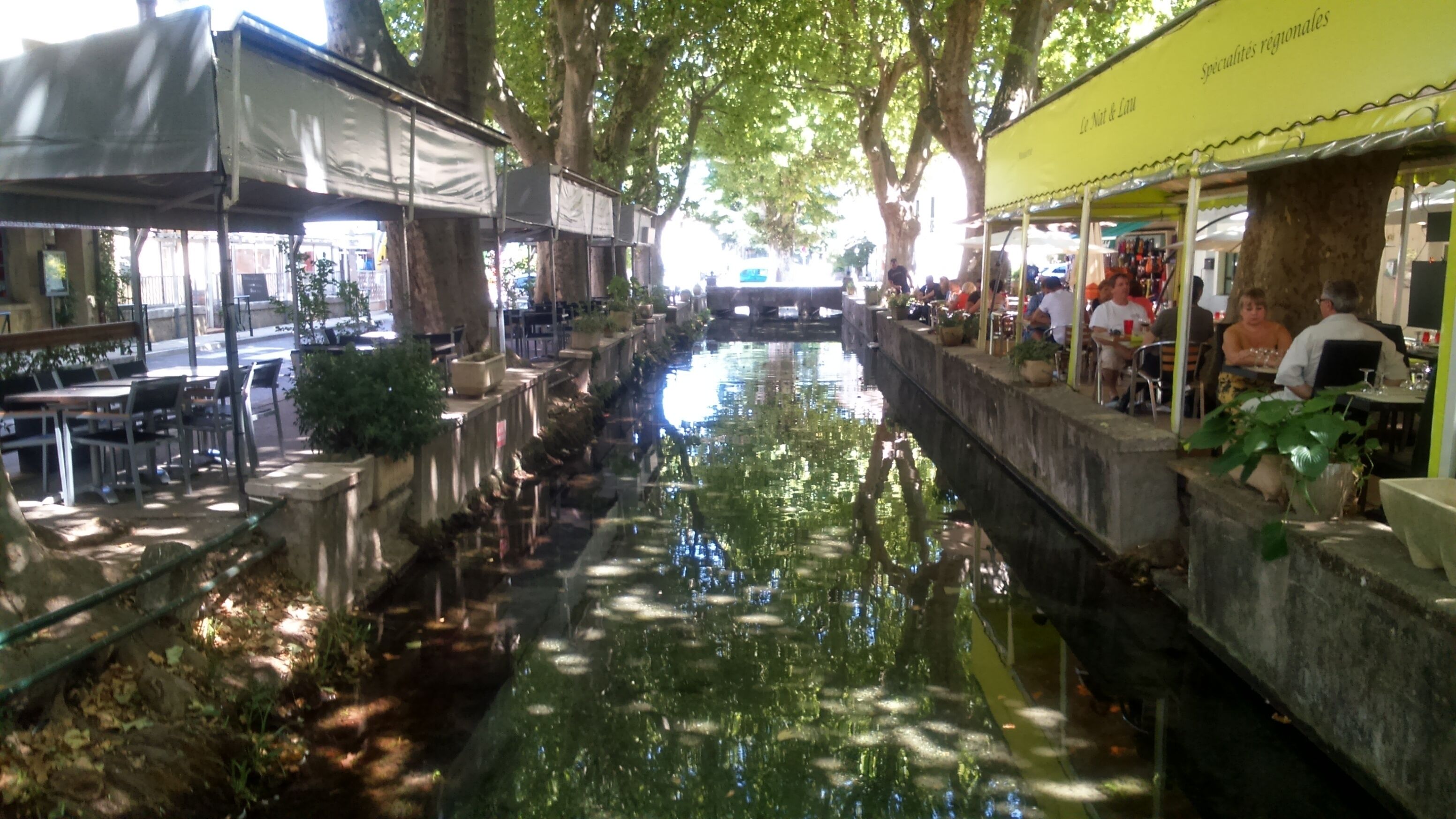 A narrow water channel with tables and chairs on either side, surrounded by trees and shaded by canopies.