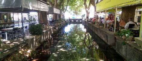 A narrow water channel with tables and chairs on either side, surrounded by trees and shaded by canopies.