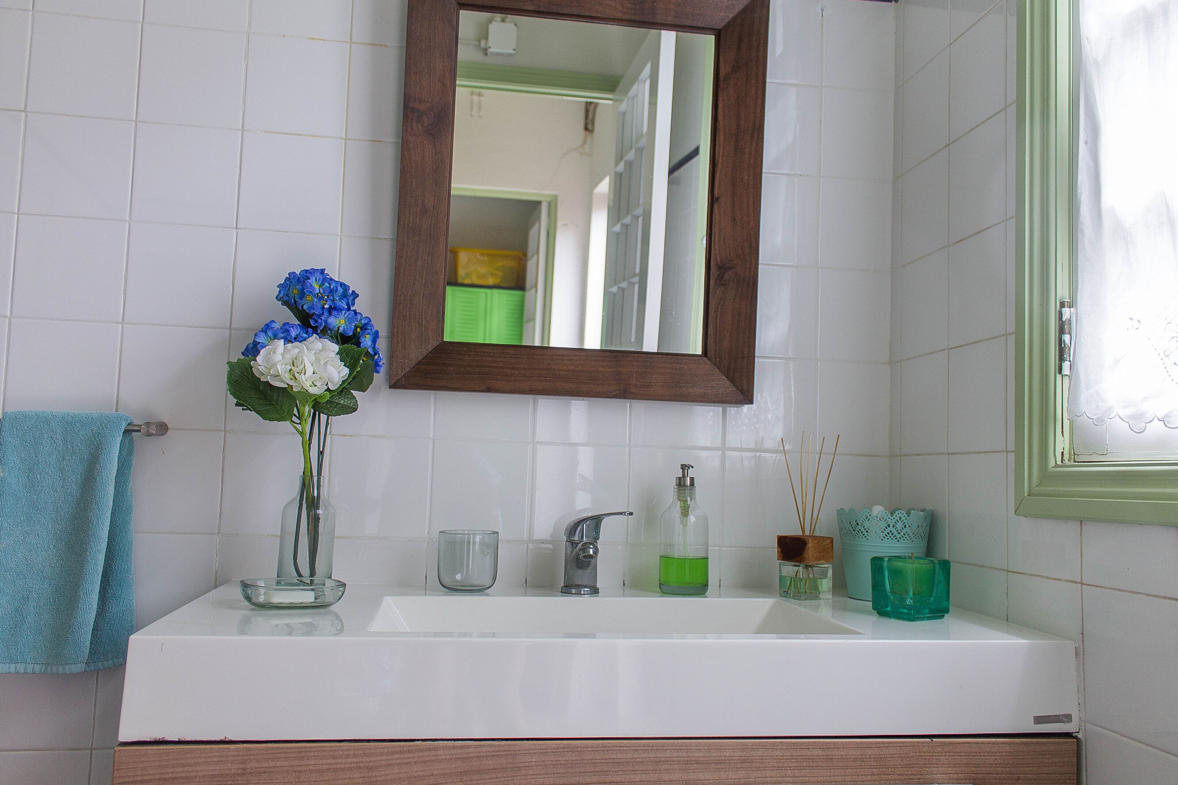 Une salle de bain avec un lavabo blanc, un miroir, une serviette bleue, un vase avec des fleurs, un distributeur de savon et une bougie.