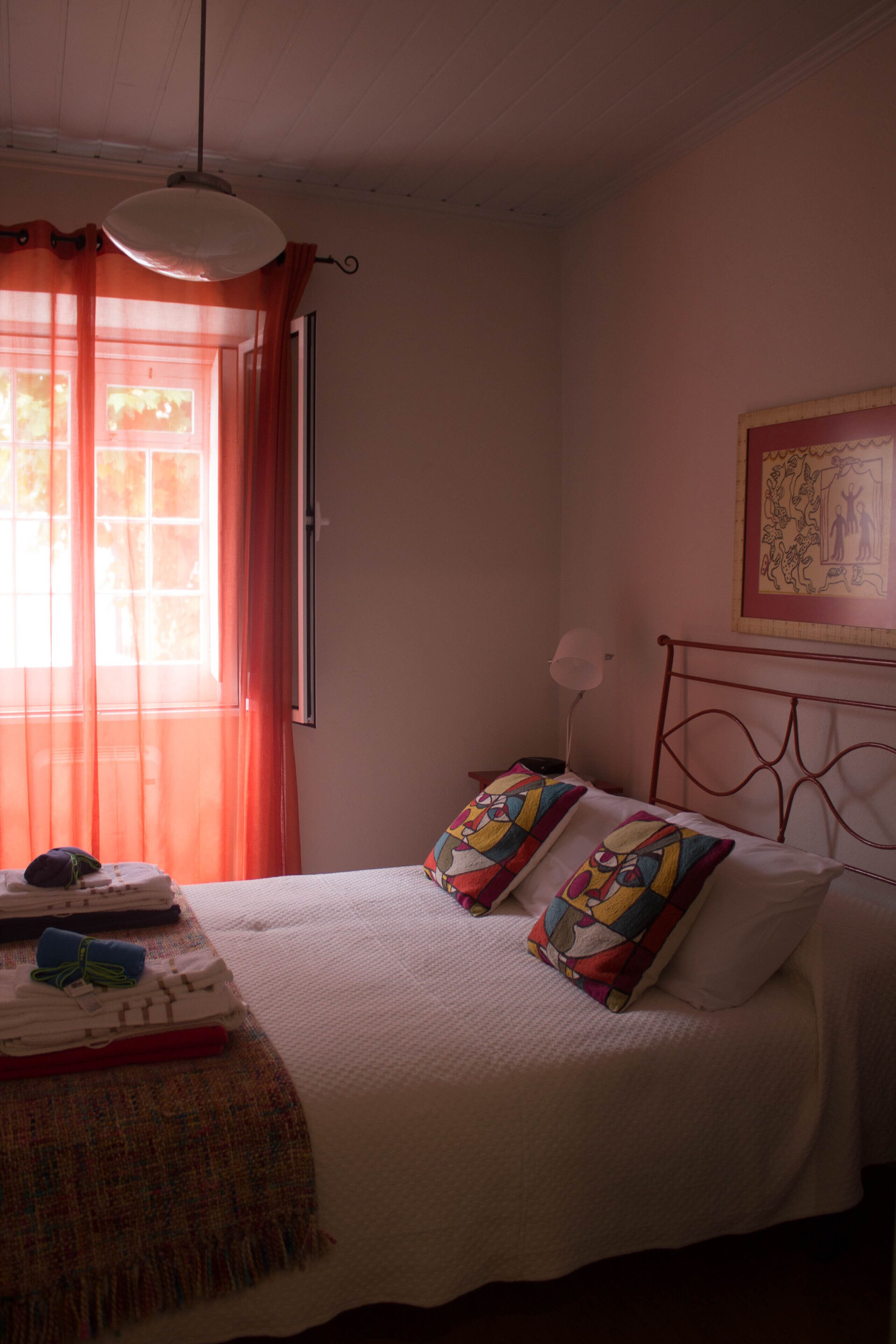 Une chambre à coucher avec un lit, une fenêtre avec des rideaux, une lampe de chevet et un tableau encadré au mur.