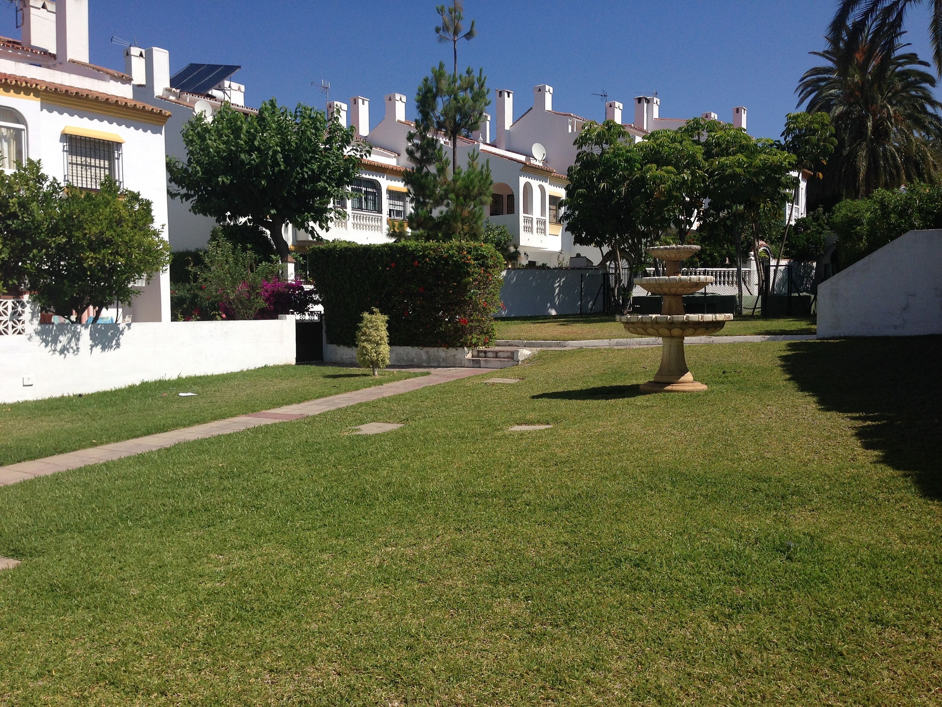 Un vecindario bien cuidado con césped, una fuente de piedra y casas blancas con balcones.
