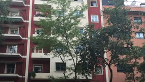 Un edificio residencial de varios pisos con balcones, un coche rojo estacionado adelante y un árbol en primer plano.