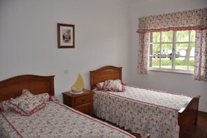 Une chambre avec deux lits simples, une table de chevet avec une lampe et une fenêtre ornée de rideaux à motifs floraux.