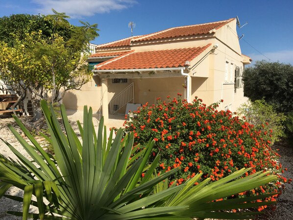 Un edificio de dos plantas con techo de tejas, un porche cubierto y un jardín con flores rojas y follaje verde.