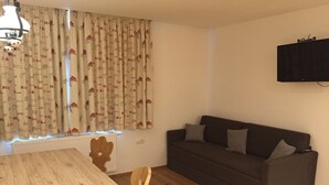 A cozy living room with a wooden table, a dark brown sofa with cushions, and a window with patterned curtains. - Convenient modern apartment in Soell, close to the ski lift (Söll)