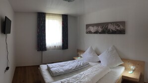 A single bed with white bedding, a mounted TV, a window with curtains, a wall-mounted picture of mountains, and a bedside table with a lamp. - Convenient modern apartment in Soell, close to the ski lift (Söll)