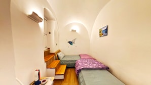 Ein schmales Zimmer mit zwei Betten, einer hölzernen Bank, einem kleinen Tisch mit Telefon und Schüssel sowie einer Wandleuchte.