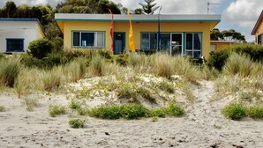 Un bâtiment à la façade jaune et aux grandes fenêtres, entouré de dunes de sable et d’une végétation clairsemée.