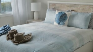 Un lit bien fait, recouvert d’une couvre-lit bleu clair, avec deux oreillers et un coussin décoratif bleu. Il y a deux serviettes sur le lit. Un tableau représentant une scène de plage est accroché au mur, au-dessus du lit.