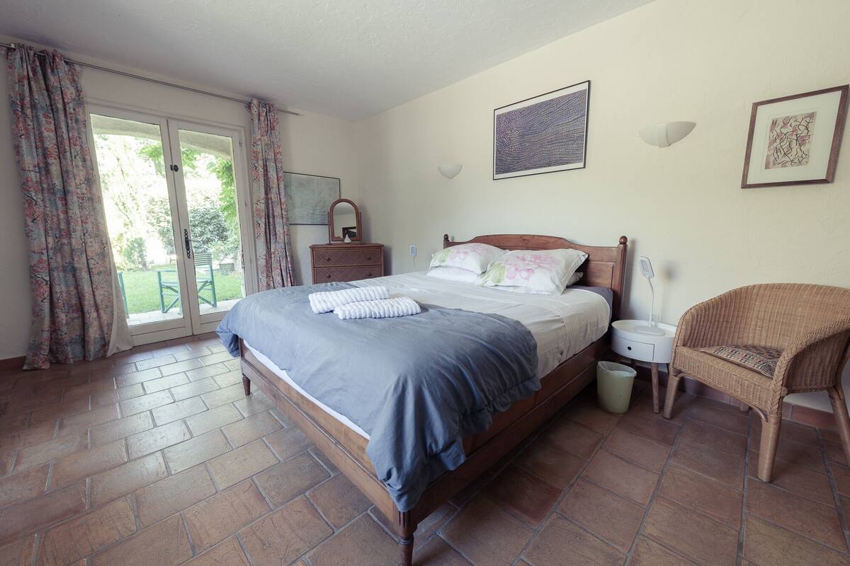 A bedroom with a wooden bed, a wicker chair, a nightstand, and a view through a glass door.