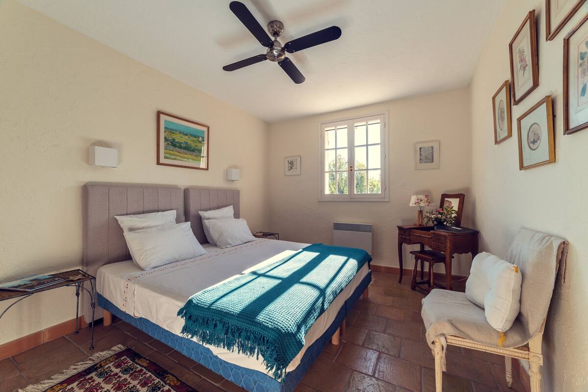 A bedroom with a bed, a desk, a chair, a ceiling fan, and a window.