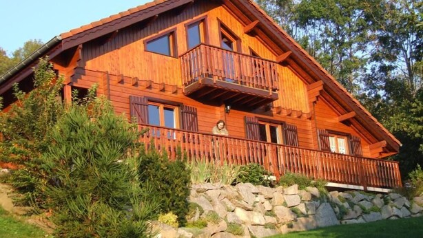 Eine Holzhütte mit einem Balkon, umgeben von Grünpflanzen und einer Steinmauer.