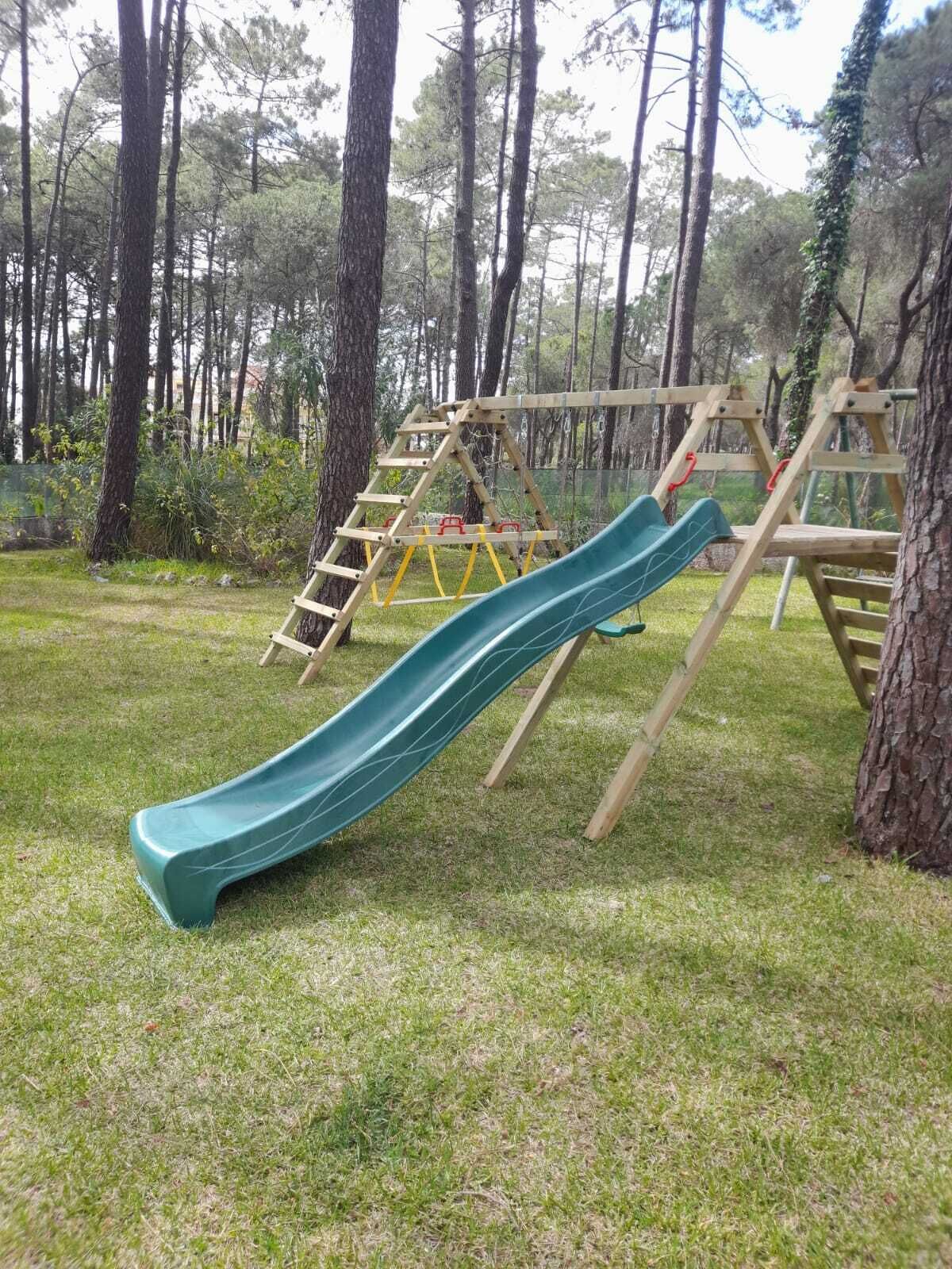 A playground with a green slide and wooden climbing structures in a grassy area surrounded by trees.