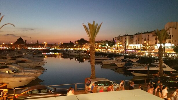 A marina at dusk with boats docked, a palm tree, and buildings in the background. - Luxury villa with large garden and pool, next to the golf course and near the beach (Vilamoura)
