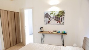 A bedroom with a bed, a wooden wardrobe, a bedside table with a kettle, cups, and a painting of a village scene. - Ailanthus Holiday Suites (Polignano a Mare)