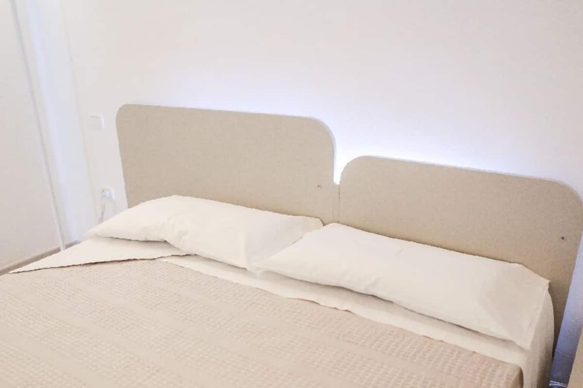 A bed with a beige headboard and two white pillows, a textured beige blanket, and a plain white wall with a small green and white sign.