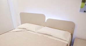 A bed with a beige headboard and two white pillows, a textured beige blanket, and a plain white wall with a small green and white sign. - Ailanthus Holiday Suites (Polignano a Mare)