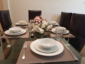 Mesa de comedor con vajilla blanca, incluyendo platos, tazones, vasos y cubiertos, rodeada de sillas tapizadas oscuras.