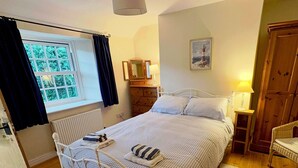 Um quarto com cama, penteadeira, mesa de cabeceira, luminária, janela com cortinas, quadro na parede e porta.