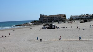 Ein Strand mit einem großen Gebäude im Hintergrund, einem Rettungsschwimmturm und Personen im Sand.