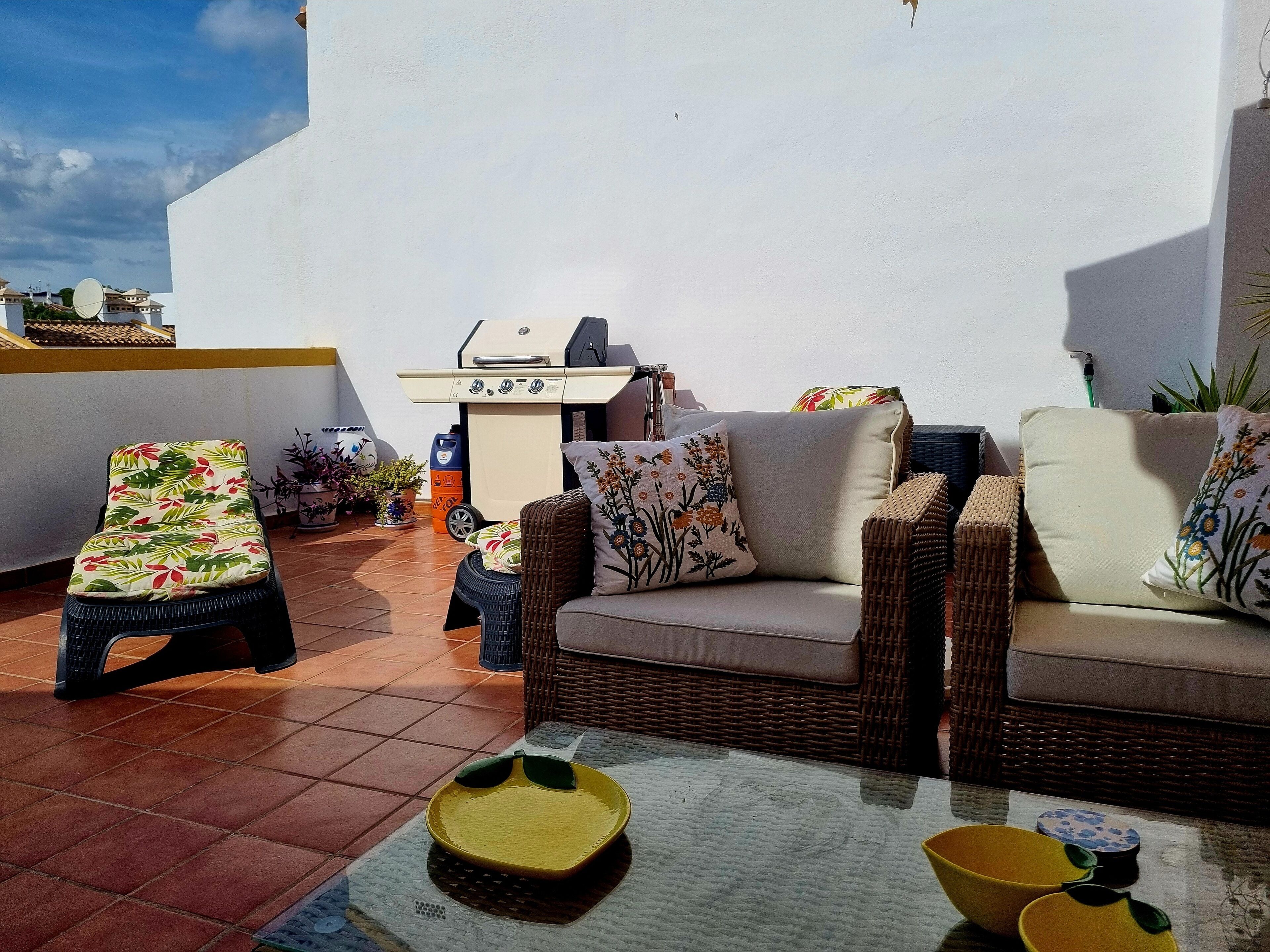 Un patio con un sofá de mimbre, una silla de lounge y una mesa de vidrio con cuencos.
