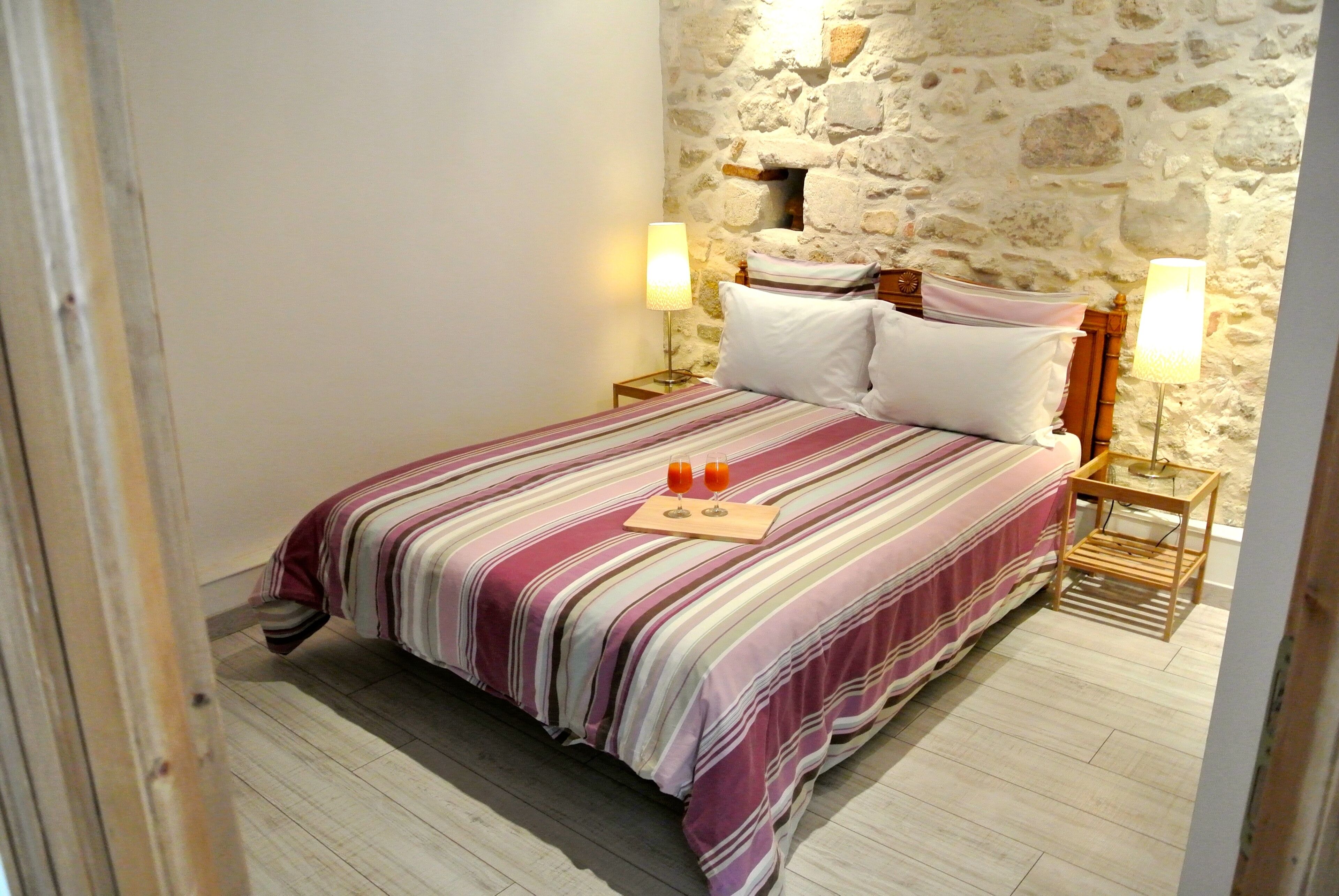 A bedroom with a stone wall, a bed with striped bedding, two bedside lamps, and a tray with two glasses.