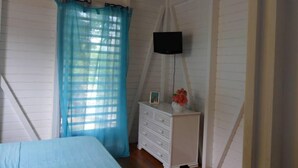 A bedroom with a bed, a white dresser, a TV mounted on the wall, and a window with blue curtains. - Gosier Charming bungalow facing the sea (Le Gosier)