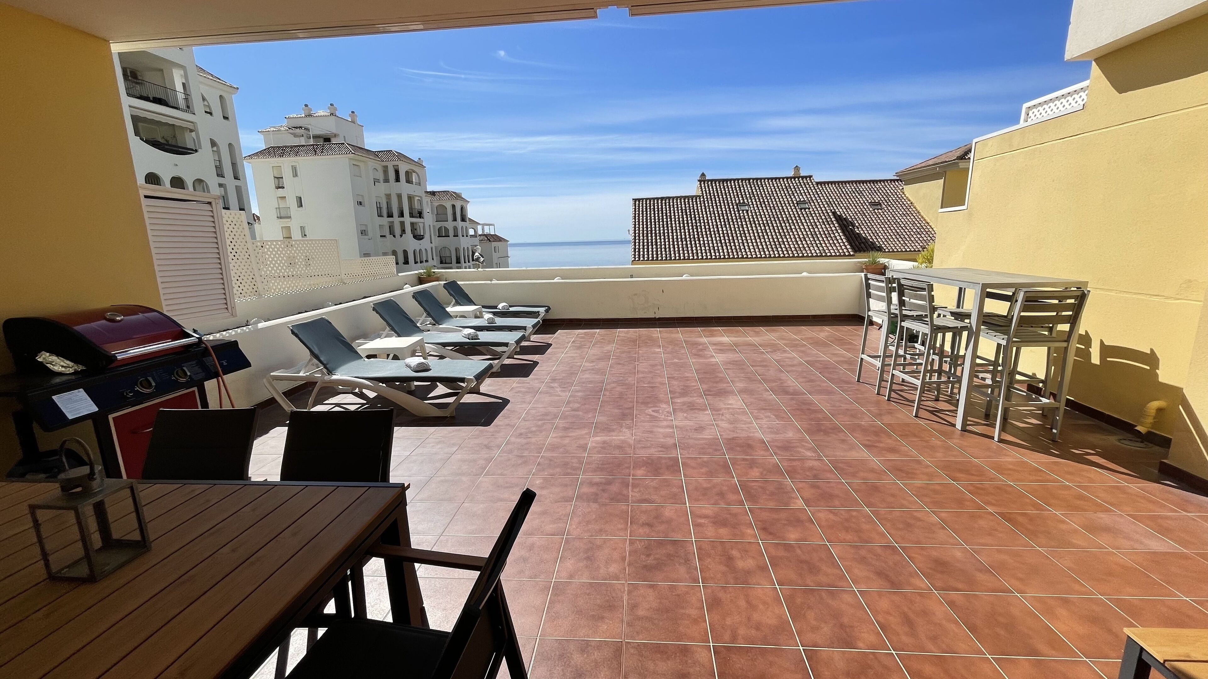 A rooftop terrace with a grill, lounge chairs, and a table.