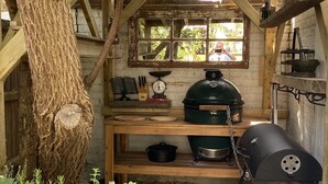 A rustic outdoor kitchen with a wooden table, a green smoker, and various cooking utensils. -  The Cook's Cottage: a coastal oasis w/outdoor kitchen, garden & cinema nr beach (Whitstable)